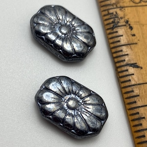 Puede incluir: Dos cuentas de metal plateadas con un diseño de flor. Las cuentas son de forma rectangular y tienen un diseño de flor en relieve en el centro.