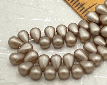 Matte pearl Czech glass drop beads in dark gold taupe, roughly fifty beads arranged on a pale background next to a wooden.
