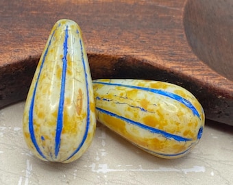 Two yellow beige Czech glass melon drop beads with blue vertical stripes, resting on a wooden dish with a white textured.