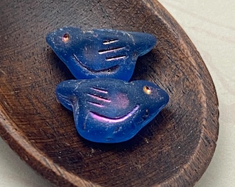 Two sapphire blue Czech glass bird beads with rainbow volcano finish in a wooden dish, showing fine white details on their.