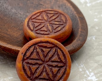Two burnt orange Flower of Life coin beads with etched surface detail, resting in a wooden dish against a pale background.