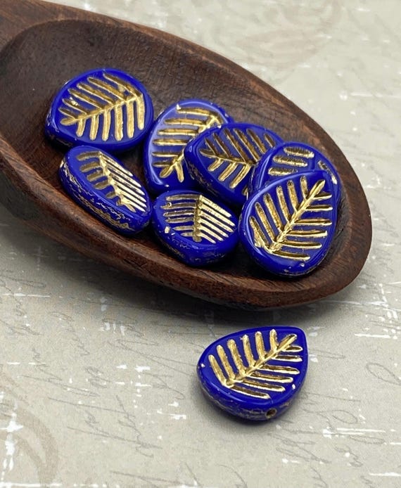 Six indigo blue Czech glass leaf beads with gold wash in a wooden dish, one separate below, showing the raised gold veining.