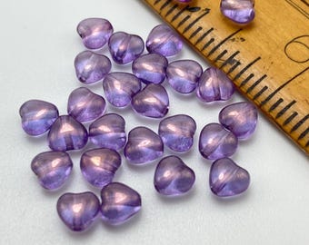 Twenty-five purple lilac AB Czech glass heart beads scattered on a white surface next to a wooden ruler for scale.