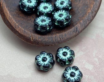 Seven black and turquoise Czech glass flower beads on a wooden dish, with three more on a pale linen surface below.