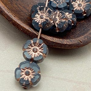 Puede incluir: Primer plano de cuentas en forma de flor azul oscuro con detalles en color cobre. Las cuentas están ensartadas en un alambre y se muestran en un cuenco de madera. Las cuentas tienen un diseño floral detallado, perfecto para la fabricación de joyas o proyectos de artesanía.