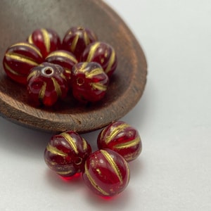 Puede incluir: Primer plano de varias cuentas de vidrio rojas con rayas doradas, descansando en una cuchara de madera. Las cuentas tienen una textura acanalada y un agujero en el centro, lo que sugiere que son para la fabricación de joyas o manualidades.