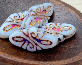 Four white Czech glass arabesque beads with gold scrollwork and rainbow volcano wash details in a wooden dish.