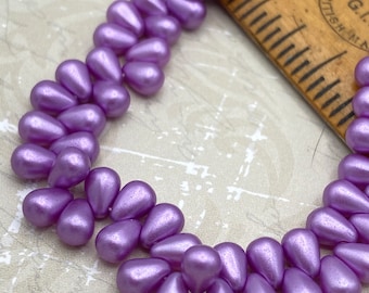 Matte pearl lilac Czech glass tiny drop beads arranged in rows next to a wooden ruler for scale.