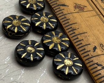 Six black Czech glass sunflower beads with gold wash details displayed on a brown wooden dish and linen cloth.