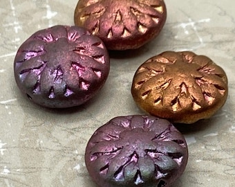 Four dahlia flower Czech glass beads in metallic purple, bronze and pink shades on a textured pale background.