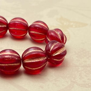 Puede incluir: Primer plano de una hilera de cuentas rojas acanaladas con detalles dorados. Las cuentas esféricas tienen un aspecto translúcido, con rayas doradas que rodean cada cuenta. Las cuentas están dispuestas en una línea curva, sugiriendo un collar o pulsera.