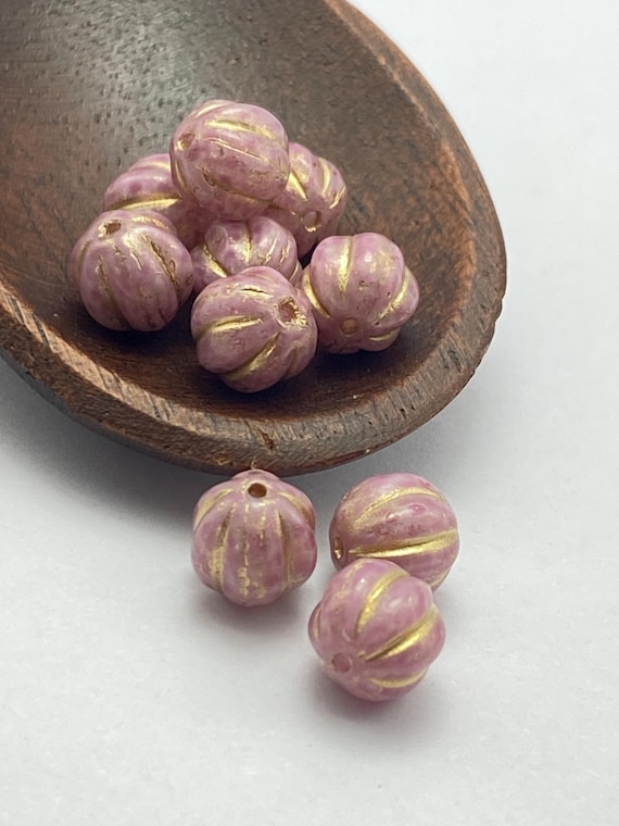 Czech glass melon beads in mauve-pink with gold wash details, arranged in a wooden dish and scattered on white surface.