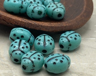 Turquoise blue ladybird beads with black spots and wing markings, some in a wooden bowl and some scattered on a pale surface.