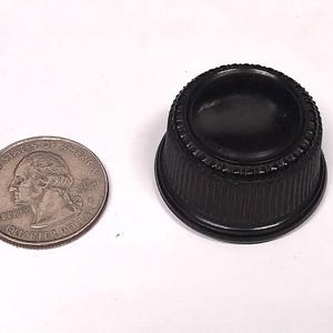 May include: A black plastic knob with a raised, textured rim and a smooth, flat top. The knob is approximately 1 inch in diameter.
