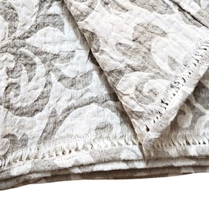 May include: A beige throw blanket with a white floral pattern. The blanket has a fringed edge.