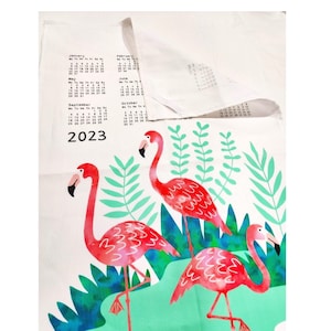 May include: A white tea towel with a calendar for 2023 and a design of three pink flamingos standing in a green tropical plant setting.
