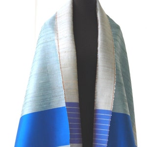 May include: A light blue and royal blue tallit with a white and gold trim. The tallit is made of silk and has a striped pattern.