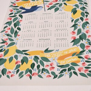 May include: A 2023 calendar with a lemon and floral theme. The calendar features a bluebird perched on a yellow banner with the text "The Zest is Yet to Come".