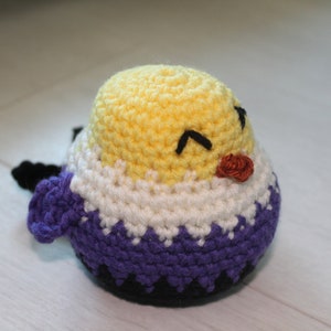 May include: A handmade crochet bird in yellow, white, purple, and black. The bird has a yellow head, white body, and purple base. It has black eyes and a small orange beak. The bird is a cute, handmade item.