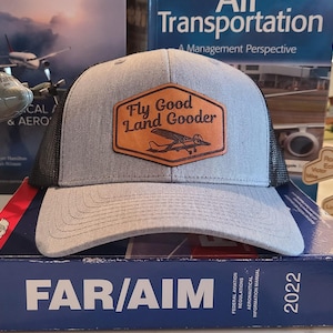 May include: A gray and black trucker hat with a brown leather patch that says "Fly Good Land Gooder" and features a small airplane illustration. The hat is sitting on top of a book titled "FAR/AIM 2022".