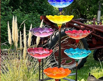 May include: A multi-tiered, outdoor bird feeder with six colorful glass bowls in shades of blue, yellow, purple, red, turquoise, and orange. The bowls are supported by a black metal frame and stand in a garden setting.