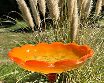 May include: An orange and yellow glass bird bath with a scalloped edge. The bird bath is mounted on a black metal stand with a decorative bronze-colored base. The background features tall ornamental grasses and greenery.