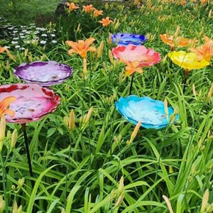 Op de afbeelding: Een verzameling kleurrijke glazen vogelbadschalen in een tuin. De schalen zijn in tinten roze, blauw, paars en geel en zijn gemonteerd op zwarte metalen palen. Oranje lelies bloeien rond de vogelbaden.