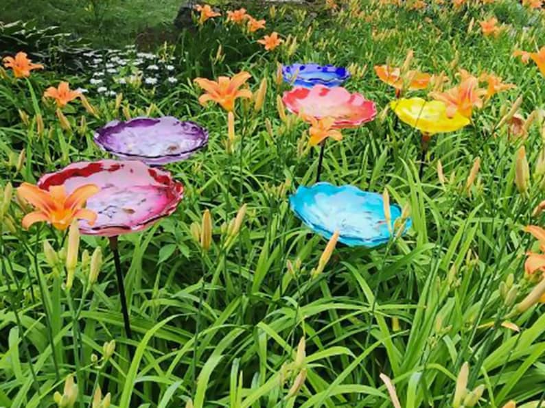 Op de afbeelding: Een verzameling kleurrijke glazen vogelbadschalen in een tuin. De schalen zijn in tinten roze, blauw, paars en geel en zijn gemonteerd op zwarte metalen palen. Oranje lelies bloeien rond de vogelbaden.