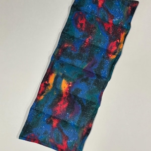 May include: A rectangular, quilted heating pad with a vibrant galaxy print. The fabric features a blend of blue, teal, red, orange, and purple hues, speckled with white dots. The pad is divided into sections by horizontal stitching.