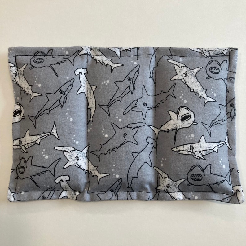 May include: A gray fabric rectangular pillow with a white shark pattern. The pillow is sewn together with a white thread. The pillow is filled with a soft material.