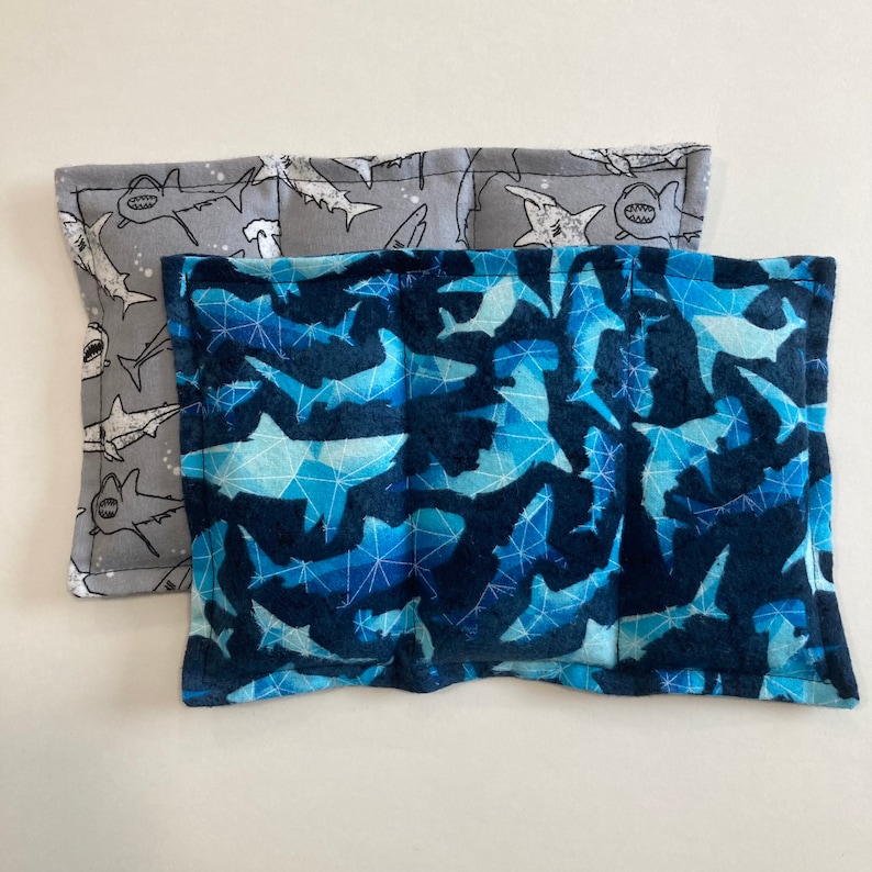 May include: Two fabric-covered heating pads with a shark pattern. The top pad is gray with white sharks, and the bottom pad is blue with white sharks.