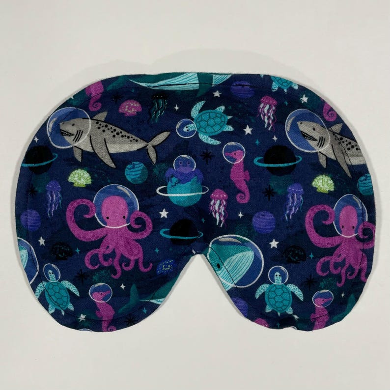 Puede incluir: Una m&aacute;scara para dormir azul con un estampado de tem&aacute;tica espacial que presenta criaturas marinas como pulpos, caballitos de mar, tiburones y tortugas. El estampado es en tonos de rosa, morado y turquesa.