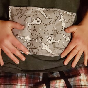 May include: A gray fabric pouch with a white shark pattern. The pouch is sewn shut with a white thread. The pouch is being held by a person's hands.