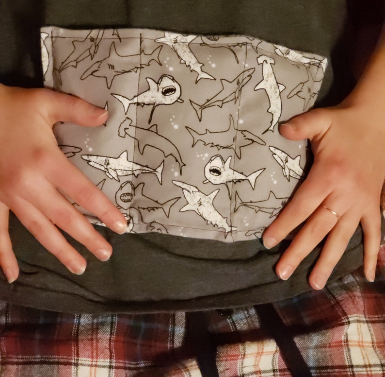 May include: A gray fabric pouch with a white shark pattern. The pouch is sewn shut with a white thread. The pouch is being held by a person's hands.