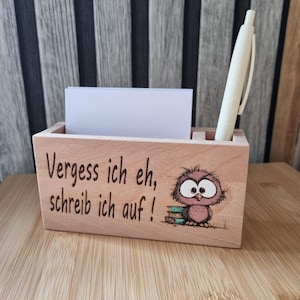 Könnte beinhalten: Ein rechteckiger Schreibtisch-Organizer aus hellem Holz mit einem Schlitz für Notizzettel und einer Halterung für einen Stift. Die Vorderseite ist mit dem Text "Vergess ich eh, schreib ich auf !" und einer kleinen lila Eule auf Büchern graviert.