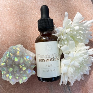 May include: A brown glass bottle of "Illuminating Essentials" Youth Facial Serum with a black dropper, white flowers, and a crystal cluster. The bottle has a white label with the product name and a moon and star design.