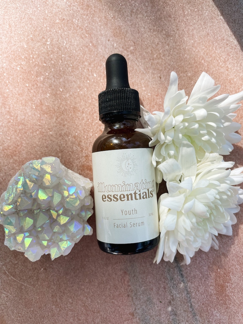 May include: A brown glass bottle of "Illuminating Essentials" Youth Facial Serum with a black dropper, white flowers, and a crystal cluster. The bottle has a white label with the product name and a moon and star design.