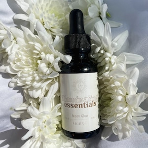 May include: A dark glass bottle of Moon Glow Facial Oil with a black dropper, surrounded by white chrysanthemum flowers. The label reads "illuminating essentials" and "Moon Glow Facial Oil". The bottle is set against a white fabric background.