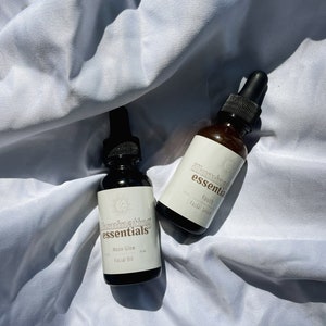 May include: Two dark amber glass bottles with black droppers, labelled "Illuminating Essentials." One reads "Moon Glow Facial Oil," the other "Youth Facial Serum." Both bottles are on a white, textured surface.