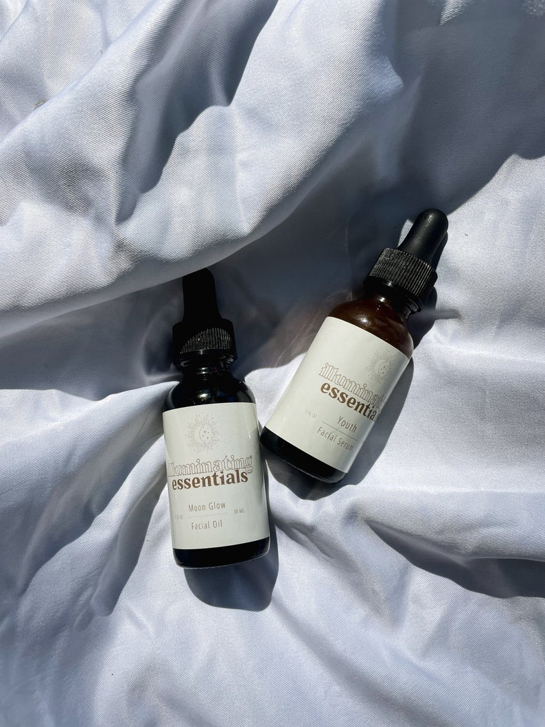 May include: Two dark amber glass bottles with black droppers, labelled "Illuminating Essentials." One reads "Moon Glow Facial Oil," the other "Youth Facial Serum." Both bottles are on a white, textured surface.