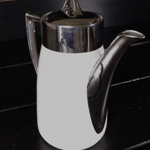 Könnte beinhalten: Eine weiße Kaffeekanne aus Keramik mit silbernem Deckel und Henkel. Die Kanne hat einen silbernen Band um die Oberseite und ein silbernes Design an der Seite.