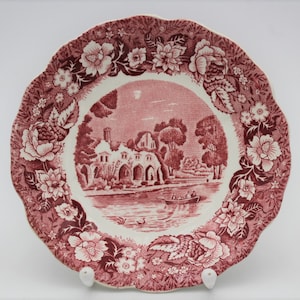 Könnte beinhalten: Ein rosa-weißer Teller aus Keramik mit einem gewellten Rand. Der Teller zeigt ein florales Design mit einem zentralen Bild eines Gebäudes und eines Sees.