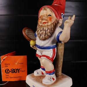 Könnte beinhalten: Eine Keramik-Wichtelfigur in Tennis-Outfit, die einen Schläger und einen Ball hält, mit roter Mütze und Bart. Die Figur steht auf einer weißen Basis mit dem Namen "TED". Ein orangefarbenes Etikett mit deutschem Text ist angebracht.