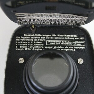 Puede incluir: Un fot&oacute;metro negro con una lente plateada y una peque&ntilde;a ventana circular negra. El fot&oacute;metro tiene una etiqueta que dice "Spezial-Notierungen f&uuml;r Kino-Kameras." e incluye instrucciones para usar el fot&oacute;metro con diferentes filtros de pel&iacute;cula.