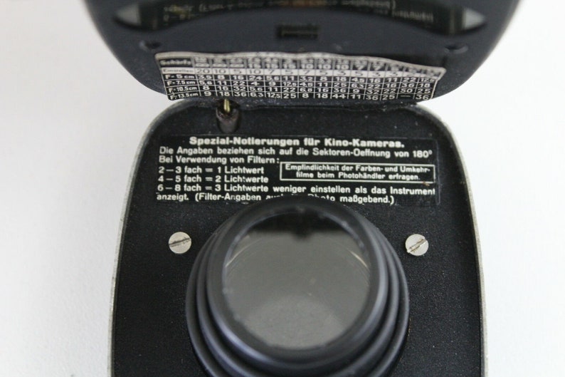 Puede incluir: Un fot&oacute;metro negro con una lente plateada y una peque&ntilde;a ventana circular negra. El fot&oacute;metro tiene una etiqueta que dice "Spezial-Notierungen f&uuml;r Kino-Kameras." e incluye instrucciones para usar el fot&oacute;metro con diferentes filtros de pel&iacute;cula.