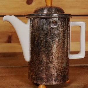 Könnte beinhalten: Eine silberne Vintage-Kaffeekanne mit weißem Ausguss und Griff. Die Kanne hat eine gehämmerten Textur und einen Deckel mit einem bernsteinfarbenen Knopf. Sie steht auf drei bernsteinfarbenen Füßen und zeigt ein klassisches Design.