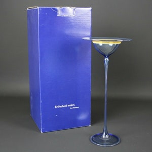 Könnte beinhalten: Eine hohe, blaue Glasvase mit einer breiten, flachen Oberseite und einem dünnen, geschwungenen Stiel. Die Vase steht auf einem blauen Karton mit dem aufgedruckten Text "Erfrischend anders. Art. Flemming".