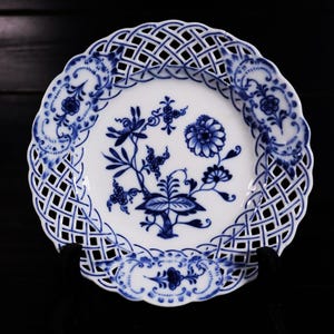 Könnte beinhalten: Weiße Porzellanteller mit blauem Blumenmuster und Gitterrand. Der Teller hat ein zentrales Blumenmotiv und aufwendige Details. Der Teller wird auf einer schwarzen Oberfläche präsentiert.