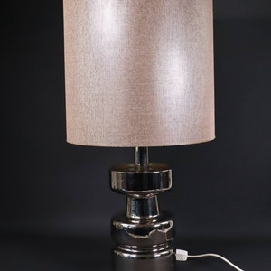 Könnte beinhalten: Eine silberne Tischlampe mit einem strukturierten, cremefarbenen Lampenschirm aus Stoff. Die Lampenbasis ist ein gestapeltes, zylindrisches Design.
