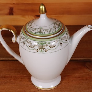 Könnte beinhalten: Eine weiße Porzellan-Teekanne mit goldener Verzierung und einem grünen und goldenen Blumendesign. Die Teekanne hat einen Deckel mit einem goldenen Knopf.